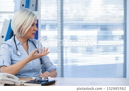 Mature female doctor gesturing and looking right at desk with stethoscope, notebook, copy space 132985562