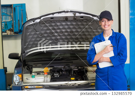 Female mechanic in blue coveralls checking open car hood in workshop holding clipboard, copy space 132985570