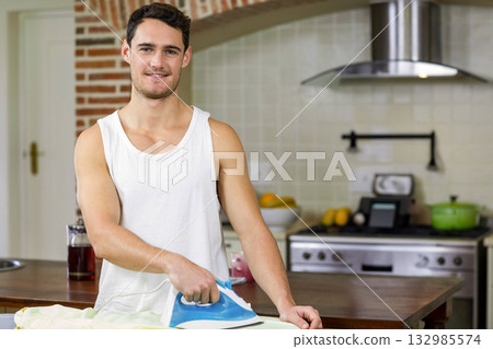 Man standing left of wooden countertop ironing garment with electric iron in kitchen, copy space 132985574