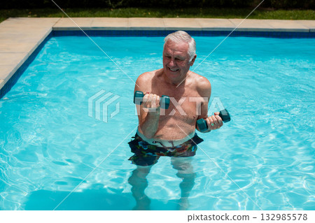 Senior man wearing swim trunks lifting dumbbells in backyard pool exercising in clear blue water 132985578