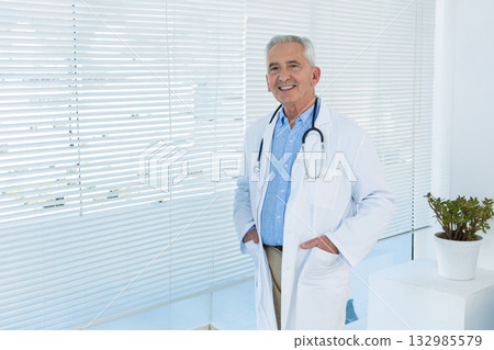 Senior doctor standing in clinic with white lab coat, stethoscope, blinds and plant, copy space Senior doctor standing in clinic with white lab coat, stethoscope, blinds and plant, copy space 132985579