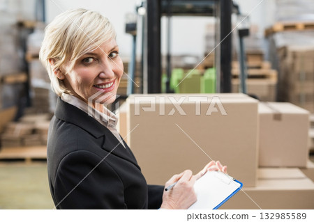 Middle-aged woman wearing blazer standing in warehouse holding clipboard near forklift, copy space Middle-aged woman wearing blazer standing in warehouse holding clipboard near forklift, copy space 132985589