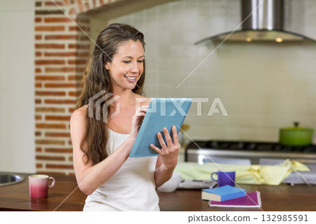 Woman using light blue tablet while standing at wooden kitchen island with mugs and books 132985591