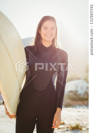 Female surfer standing on beach holding surfboard beside coastal house in wetsuit under backlight Female surfer standing on beach holding surfboard beside coastal house in wetsuit under backlight 132985592
