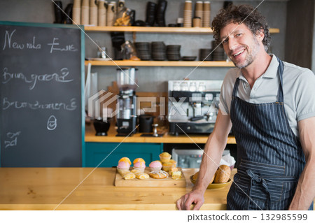 Male barista wearing apron leaning on cafe counter showing pastries, chalkboard menu, copy space Male barista wearing apron leaning on cafe counter showing pastries, chalkboard menu, copy space 132985599
