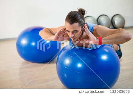 Man in his twenties balancing on blue exercise ball with metallic gym balls at fitness studio 132985603