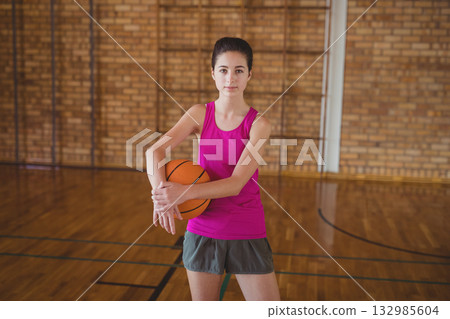 Teenage female athlete holding orange basketball standing center court wearing pink top gray shorts Teenage female athlete holding orange basketball standing center court wearing pink top gray shorts 132985604
