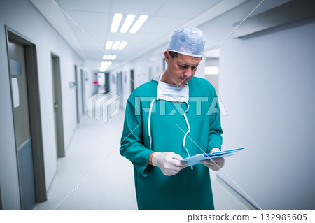 Mid adult man standing reading charts in hospital corridor wearing scrubs, cap, mask and gloves Mid adult man standing reading charts in hospital corridor wearing scrubs, cap, mask and gloves 132985605