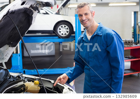 Male mechanic in blue coveralls pointing at coolant reservoir under hood in repair shop, copy space 132985609