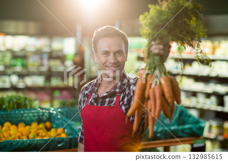 Male produce clerk smiling and presenting fresh carrots beside mesh orange bins at supermarket Male produce clerk smiling and presenting fresh carrots beside mesh orange bins at supermarket 132985615