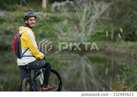 Man smiling astride mountain bike at forest pond edge wearing rain jacket and helmet, copy space Man smiling astride mountain bike at forest pond edge wearing rain jacket and helmet, copy space 132985623