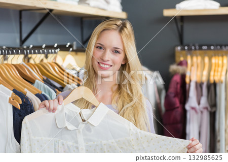 Woman holding light-colored blouse on hanger inside clothing store among clothing racks and shelves 132985625
