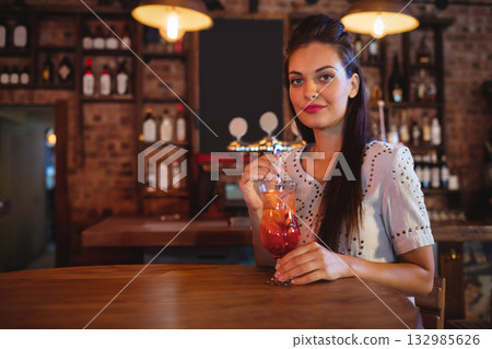 Woman in twenties holding orange-red cocktail with straw at bar counter with brick wall, copy space Woman in twenties holding orange-red cocktail with straw at bar counter with brick wall, copy space 132985626