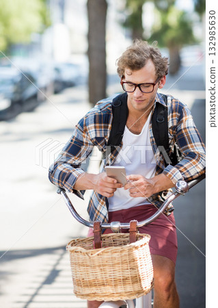 Man cycling with backpack and wicker basket along tree-lined sidewalk while checking smartphone 132985630