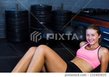 Woman leaning back on hands with bent knees on rubber mat near medicine balls and dumbbells Woman leaning back on hands with bent knees on rubber mat near medicine balls and dumbbells 132985635