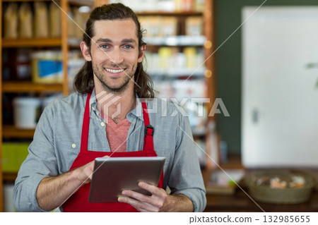 Male store clerk wearing red apron, holding digital tablet, checking inventory in shop aisle 132985655
