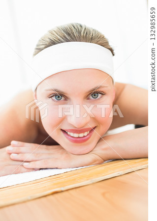 Female client lying on white towel covering wooden frame massage table in spa room wearing headband 132985659
