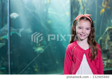 School-age girl wearing lace dress and floral headband watching fish by aquarium, copy space 132985660