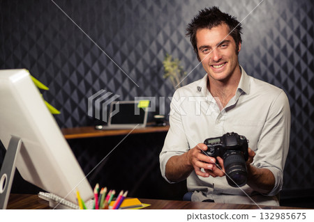 Adult man sitting at desk in studio holding DSLR camera near monitor and pencils, copy space Adult man sitting at desk in studio holding DSLR camera near monitor and pencils, copy space 132985675