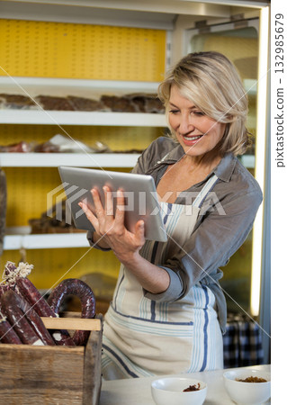 Female shopkeeper wearing striped apron holding tablet while reviewing stock at meat counter Female shopkeeper wearing striped apron holding tablet while reviewing stock at meat counter 132985679
