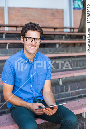 Man wearing black glasses and blue shirt sitting on schoolyard bleachers holding tablet Man wearing black glasses and blue shirt sitting on schoolyard bleachers holding tablet 132985680