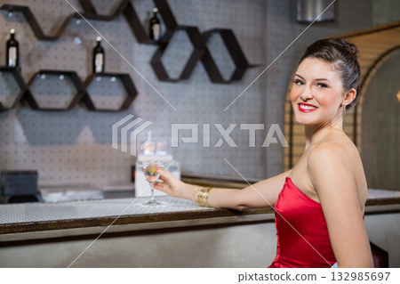 Woman in fitted red cocktail dress leaning on bar counter while holding stemmed glass in lounge 132985697