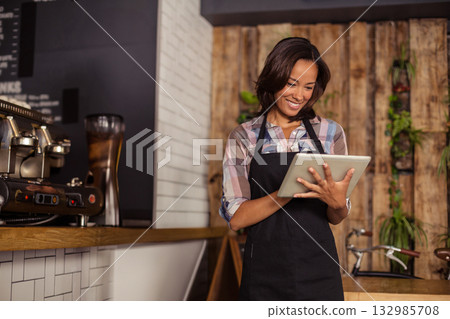 Asian woman barista wearing apron using tablet at tile counter near espresso and plants, copy space 132985708