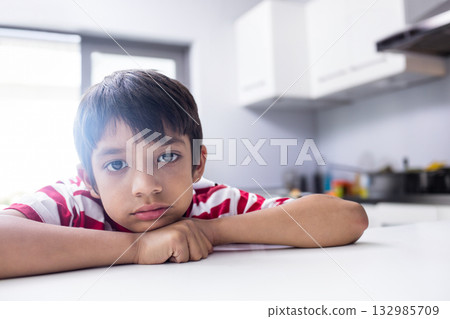 Indian school-age boy wearing striped T-shirt leaning on countertop near window in kitchen 132985709