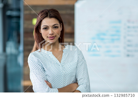 Female professional standing with arms crossed in office with whiteboard and glass partition Female professional standing with arms crossed in office with whiteboard and glass partition 132985710