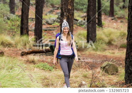 Woman in her 20s walking through pine forest clearing carrying purple backpack and wearing knit hat Woman in her 20s walking through pine forest clearing carrying purple backpack and wearing knit hat 132985723