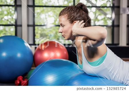 Female athlete balancing on blue exercise ball in fitness studio holding head beside red dumbbells 132985724