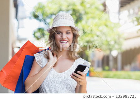 Woman standing on sunny retail street wearing sun hat, holding shopping bags and smartphone 132985725