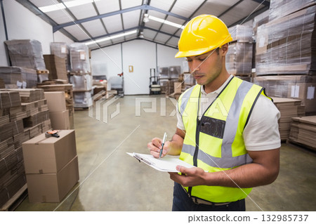 Man in hard hat and vest checking boxes using clipboard in warehouse aisle, copy space 132985737
