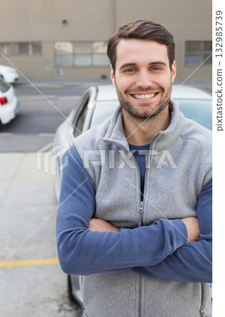 Male crossing arms and standing in parking lot in front of silver sedan near building windows 132985739