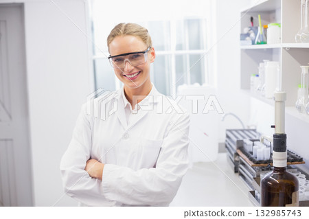 Female scientist smiling while standing in bright lab by bench with pipettes wearing white coat Female scientist smiling while standing in bright lab by bench with pipettes wearing white coat 132985743