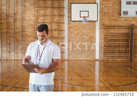 Mature adult male coach holding clipboard with pen near basketball hoop and wall bars in gymnasium 132985758