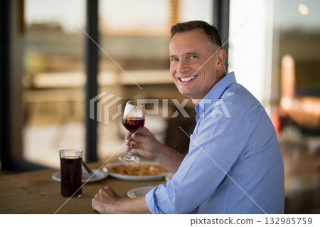 Middle-aged man holding red wine glass, smiling at wooden restaurant table with pasta and dark soda Middle-aged man holding red wine glass, smiling at wooden restaurant table with pasta and dark soda 132985759