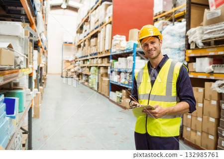 Male worker wearing hard hat inspecting boxes in warehouse aisle with clipboard, copy space Male worker wearing hard hat inspecting boxes in warehouse aisle with clipboard, copy space 132985761