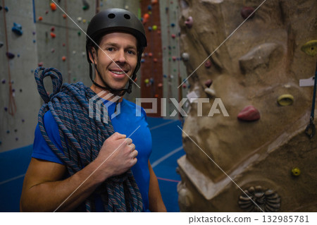 Mid adult man standing in climbing gym wearing black helmet holding rope beside colorful holds Mid adult man standing in climbing gym wearing black helmet holding rope beside colorful holds 132985781