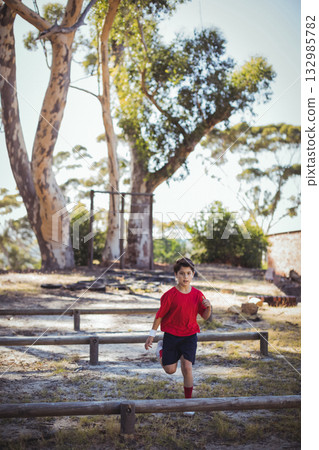 Boy running along dirt clearing navigating wooden-beam obstacle course holding oval ball in park Boy running along dirt clearing navigating wooden-beam obstacle course holding oval ball in park 132985782