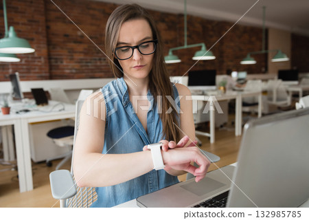 Woman office worker checking smartwatch on wrist while sitting at laptop in open plan office Woman office worker checking smartwatch on wrist while sitting at laptop in open plan office 132985785