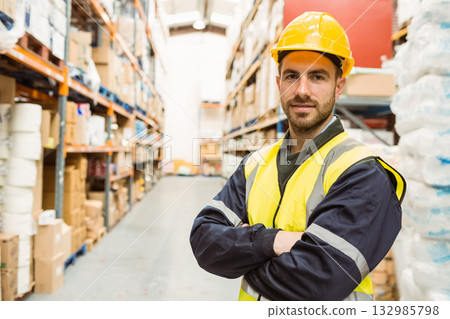 Man wearing yellow hard hat, reflective vest standing in warehouse aisle among boxes, copy space 132985798