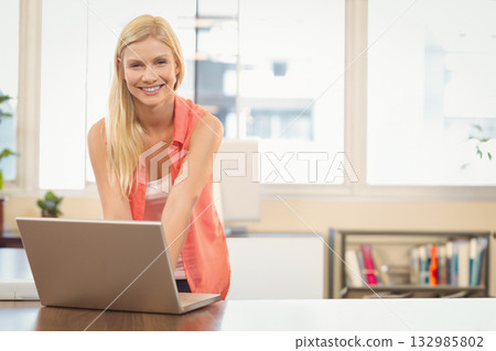 Woman leaning over wooden desk, working on laptop in modern office by potted plant and bookshelf Woman leaning over wooden desk, working on laptop in modern office by potted plant and bookshelf 132985802