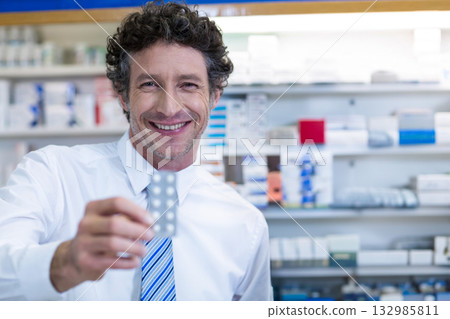 Smiling male pharmacist standing behind counter with medication shelves, offering pill blister pack Smiling male pharmacist standing behind counter with medication shelves, offering pill blister pack 132985811