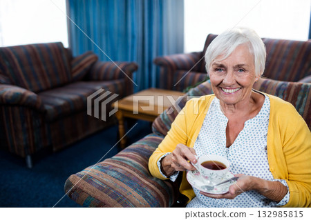 Senior woman holding floral teacup and saucer while sitting in striped armchair at home, copy space Senior woman holding floral teacup and saucer while sitting in striped armchair at home, copy space 132985815