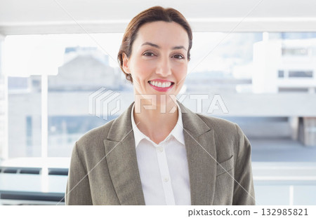 Businesswoman standing and smiling in modern office in front of large windows and radiator Businesswoman standing and smiling in modern office in front of large windows and radiator 132985821