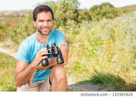 Man kneeling on dirt path in grassy field holding binoculars and smiling toward camera, copy space 132985826