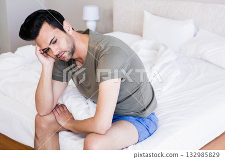 Mid adult man sitting on edge of unmade bed in lit bedroom, holding head beside lamp 132985829