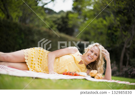 Woman reclining wearing yellow dress on striped picnic blanket in park with croissants and tomatoes Woman reclining wearing yellow dress on striped picnic blanket in park with croissants and tomatoes 132985830