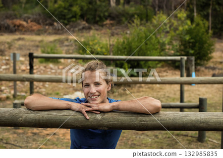 Fit woman wearing blue athletic shirt leaning on wooden rail at obstacle course with dirt patches 132985835